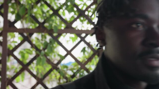 Camera view shows dreadlocks man backing crisscross iron grille, x pattern filling frame, head turning left then right, tensed yet curious mood, metal lattice forming bold graphic texture