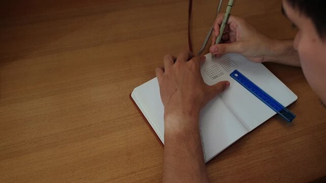 Static shot of male student writing lines for a calendar view inside a bullet journal over wooden desk.