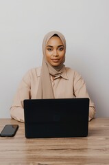 A confident and focused individual, professionally dressed and wearing a modest head covering, sits attentively at a polished wooden desk. With a modern laptop open in front and a smartphone placed ne