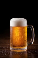 A frosty glass mug filled with a rich, golden beverage, crowned with a thick layer of creamy white foam. Numerous condensation droplets cling to the chilled glass surface, cascading onto the dark, ref