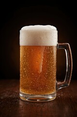 A perfectly poured mug of cold, golden amber beverage stands prominently on a dark, rustic wooden surface. The glass is covered in glistening condensation, emphasizing its chilled temperature and refr
