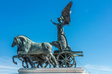 View of bronze horses pulling a chariot carrying a winged figure, all against a brilliant blue sky, Rome, Italy.