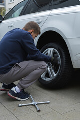 Man changing car tire using electric drill and jack on city sidewalk. DIY auto repair, maintenance, seasonal tire change, mechanical work, vehicle service, urban life.