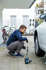 Man changing car tire using electric drill and jack on city sidewalk. DIY auto repair, maintenance, seasonal tire change, mechanical work, vehicle service, urban life.