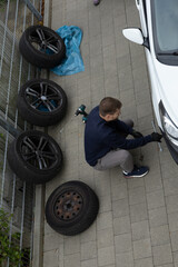Man changing car tire using electric drill and jack on city sidewalk. DIY auto repair, maintenance, seasonal tire change, mechanical work, vehicle service, urban life.