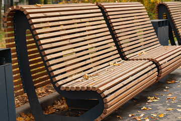 Modern wooden bench in a park with autumn leaves scattered on the ground. The bench features a sleek design with horizontal slats and a sturdy frame.