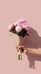 Enchanting Bouquet in Hand: A tender hand delicately presents a bouquet of vibrant, pink peonies against a backdrop of soft pastel hue, creating a scene of understated elegance and floral beauty.