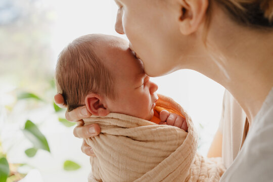 Beautiful mother kissing her baby. Authentic photography of mother holding her newborn baby after labor. Newborn in mother's hands. Baby care. Childbirth and motherhood concept.