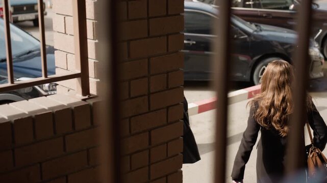 Two students wearing long winter jackets meet at hallway entrance, walking opposite directions past block rails, sunlight on hair, parked cars beyond and candid campus vibe with gentle breeze