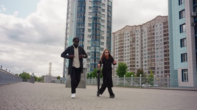 front angle shot two friends walking with boldness on modern city promenade near tall buildings, Confident stride under cloudy sky, ground level highlights urban style and relaxed conversation energy