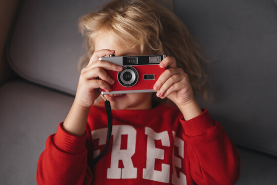 A blond, curly-haired girl holds a red retro camera while lying on a sofa. Children's fascination with retro toys.