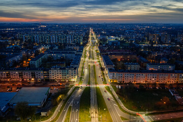 Aerial night cityscape with illuminated streets and buildings seen from drone, glowing lights of urban infrastructure and sports field creating modern metropolitan atmosphere at sunset and dusk