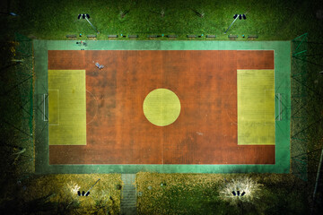 Aerial view of illuminated sports field at night, glowing green football pitch surrounded by dark city background, stadium floodlights creating geometric symmetry and modern urban atmosphere