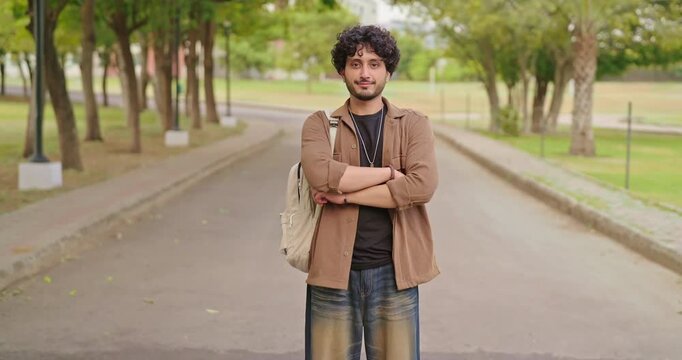 Indian happy young adult gen z man male look camera cross arm standing outdoor area. Smart cool proud pupil boy guy wear bag fold hand staring cam do casual pose enjoy new day outside place