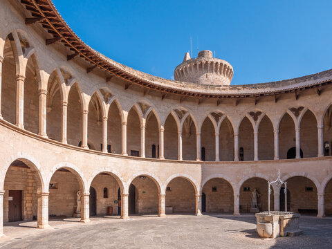 Castell de Bellver, Palma de Mallorca, historic circular cloister with arched colonnades and a central fountain stands in a courtyard under a clear blue sky, Majorca