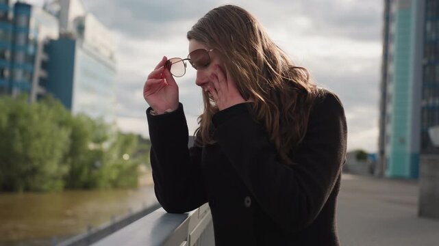Tourist wearing thick jacket covers face in discomfort while standing by river, sunlight reflecting on glasses as skin turns red, expressing sensitivity and tension amid calm modern architecture