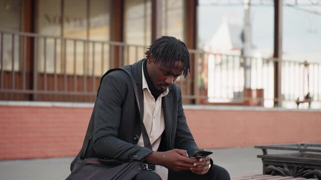 Dreadlocked man with short beard wearing layered clothing sits on wooden bench using smartphone outdoors, showing calm focus and stylish appearance in modern city with soft daylight reflections