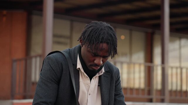 Close view of dreadlocked man with rough twisted natural hair looking down at mobile phone, dressed in layered clothing, showing style, and natural expression in soft daylight urban setting
