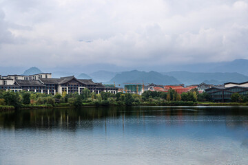 Obraz premium Cityscape featuring modern buildings with traditional Asian architectural roofs beside a calm lake, surrounded by lush greenery and misty mountains under cloudy skies