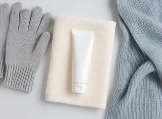 Winter hand care concept. Moisturizing cream tube on soft towel with knitted gloves and scarf. Minimal composition in cozy pastel colors for cold season skincare theme.