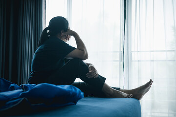 A woman sitting alone on bed facing window in dark room, symbolizing depression, loneliness, anxiety, sadness, mental health struggle, insomnia and emotional stress in life challenges.