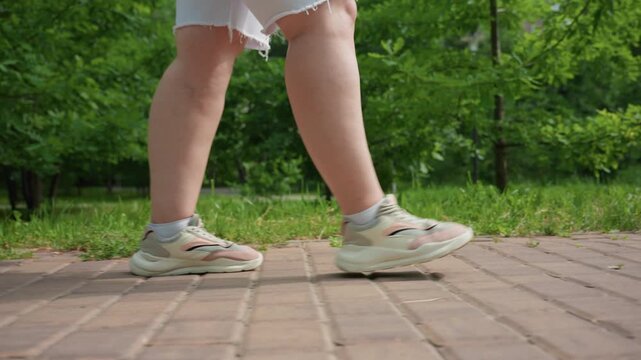 side view of fat lady strolling along brick path surrounded by lush green trees and grass during bright summer day, wearing sneakers and white shorts, enjoying peaceful outdoor walk