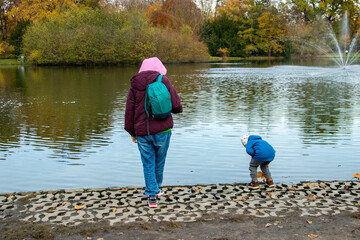 A woman and child by the lake on a cloudy autumn day. The child leans toward the water, and a fountain sprays in the distance.