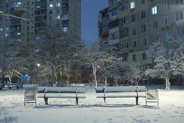 A snowy city park with benches and streetlights at night.