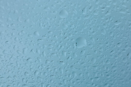 A macro, high-detail view of countless water droplets and streaks on a cool blue glass surface, creating a beautiful, abstract texture ideal for a wet weather background.