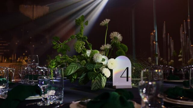 Luxury wedding reception. Festive table setting with flower arrangement, number 4 for guests on black tablecloth, green napkins, candles on backdrop, soft moving searchlight rays in hall restaurant.