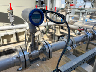 Naklejka premium Ultrasonic flow meter mounted on industrial pipeline with digital display. Close-up of metallic pipe, flanges, bolts and connected cable. Background shows additional valves and piping.