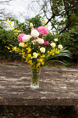 Vibrant bouquet of white roses, pink carnations, and yellow filler flowers in a glass vase, on a textured, rustic wooden table. Outdoor scene captured in daylight. Symbolizes love, celebration.