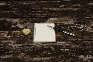 Top view of a closed antique gold-tone pocket watch, a lined notebook, and a metal fountain pen on a weathered, rustic wooden table. Symbolizes writing, planning, time, and history.