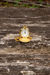 Vintage gold pocket watch with open cover and chain, resting on a textured, weathered rustic wooden table. Outdoor setting. Symbolizes time, luxury, history, and nostalgia.