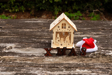 Christmas scene: Miniature wooden house, XMAS letters, and Santa hat on rustic wooden table. Ample copy space left. Outdoor in daylight. Symbolizes holidays, home, and winter decor.