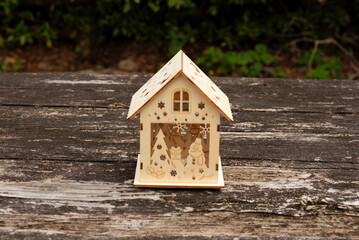 Miniature wooden house with intricate winter scene cutout design, resting on a weathered, rustic wooden table. Outdoor setting. Symbolizes Christmas, home, winter decor, and cozy holidays.
