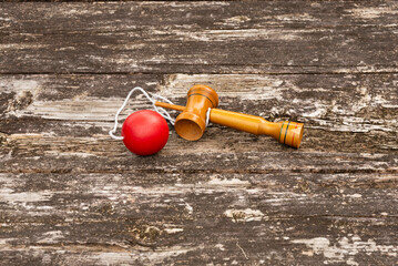 Single traditional Japanese cup-and-ball toy (kendama) with red ball and string, displayed on a weathered rustic wooden table. Outdoor. Symbolizes traditional games, skill, and Japanese culture.
