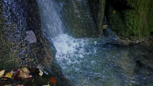 Small waterfall cascading over moss-covered rocks in slow motion, with vibrant autumn leaves creating a peaceful natural scene