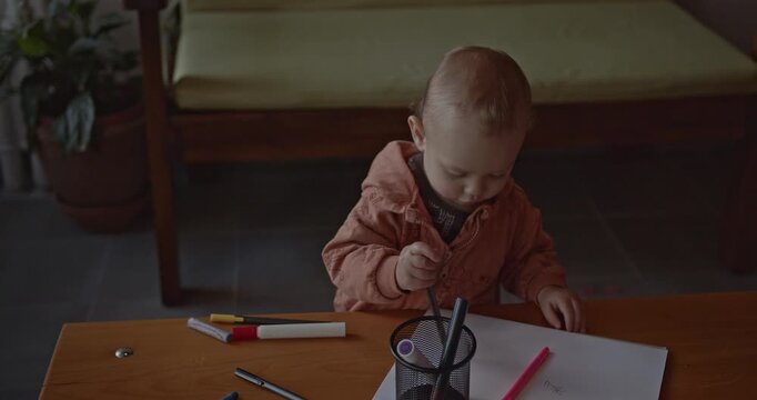 A curious baby explores colorful markers and paper, scribbling and playing at a wooden table in a cozy home setting.