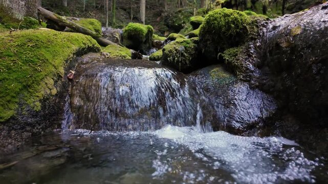 Small mountain waterfall flowing between mossy rocks in slow motion, crystal water creating relaxing natural sound and movement