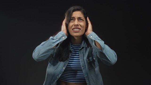 Woman covering ears with hands in studio against black backdrop wearing striped top and denim jacket; sensory overload distress.