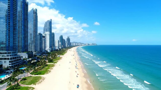 Aerial view of a bustling city beach with highrise buildings and palm trees lining the shore under a clear blue sky