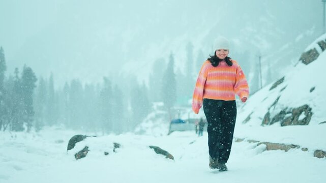 4K Shot of happy Indian woman walking towards camera while enjoying snowfall in mountains at Sonamarg, Jammu and Kashmir, India. Travel, tourism and holidays concept. Snowfall during winter.
