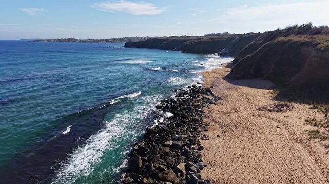 At a quiet beach, sunlit rocks border the soft sand, where waves playfully lap the shore and the horizon stretches far beyond. Peace surrounds the tranquil scene.