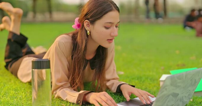 Indian young adult gen z busy female use laptop look screen check online new e work skill in fresh air park. Close up of happy girl lying on grass do study read book turn pages spend break time place