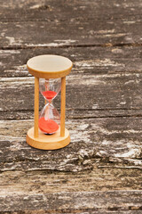 Wooden hourglass with red sand running through, standing on a textured, weathered rustic wooden table. Outdoor setting. Symbolizes time, passage, countdown, and deadline.