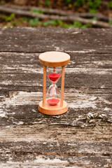 Wooden hourglass with red sand running through, standing on a textured, weathered rustic wooden table. Outdoor setting. Symbolizes time, passage, countdown, and deadline.