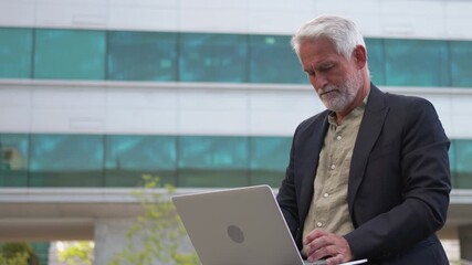 Confident senior businessman using laptop outdoors near office building, working remotely in modern urban environment during daytime - Powered by Adobe