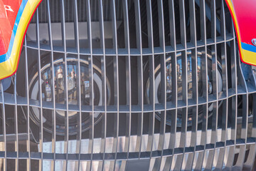 Close-up of a colored grill with vertical bars and horizontal sections, angled view of vehicle headlights, shallow depth of field, natural light, red, black, white