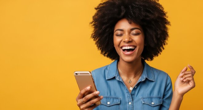 A happy young black woman with an afro hairstyle laughing while looking at her smartphone against a vibrant yellow background
